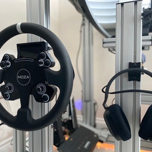 May include: A black racing steering wheel with a white stripe and the word "MOZA" on it. The steering wheel is mounted on a silver metal frame. A pair of black headphones are hanging on a silver metal frame.