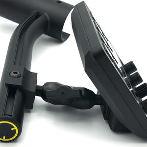 May include: Black electronic device with multiple knobs and buttons, mounted on a curved black support arm. The support arm has a yellow and black circular design on the end. The device is angled, showing its side profile.