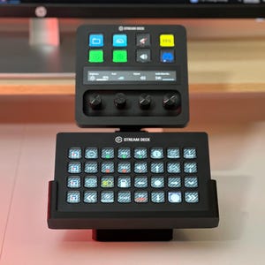 May include: A black Stream Deck device with multiple buttons and a small screen. The device has a top section with a screen and knobs, and a bottom section with numerous illuminated buttons. The Stream Deck logo is visible.