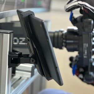 May include: A black tablet monitor mounted on a silver metal stand. The tablet is angled towards the right and has a black screen. The stand is attached to a larger silver metal structure.