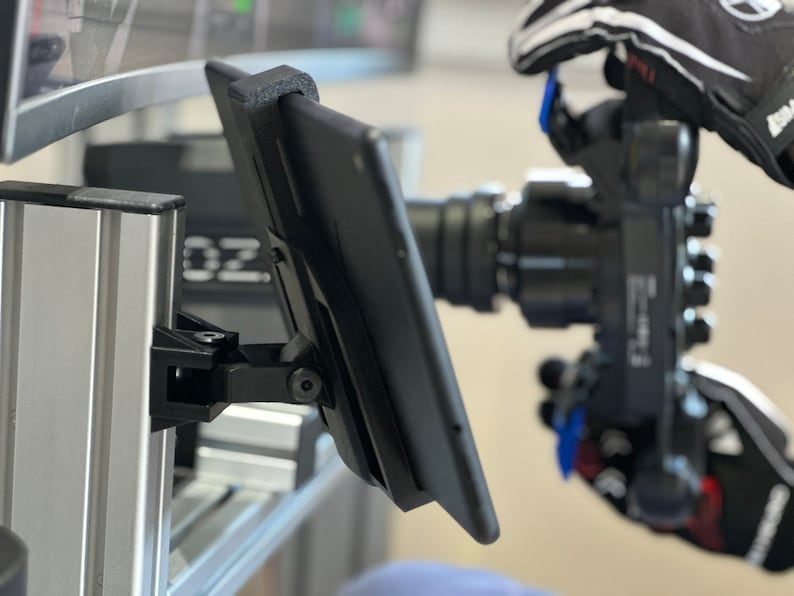 May include: A black tablet monitor mounted on a silver metal stand. The tablet is angled towards the right and has a black screen. The stand is attached to a larger silver metal structure.