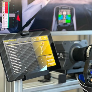 May include: A black tablet displaying a racing leaderboard with the text "N&Uuml;RBURGRING RACE" and a map of the racetrack. The tablet is mounted on a stand and is positioned in front of a racing simulator.