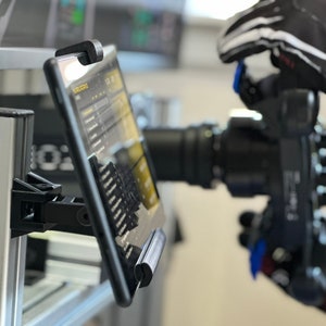 Puede incluir: Una tableta negra con una pantalla que muestra un menú de opciones está montada en un poste plateado con una abrazadera negra. La tableta está posicionada a la izquierda de una cámara negra con una lente grande.