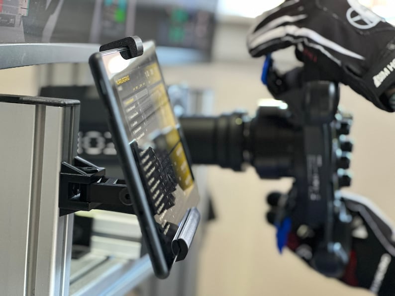 May include: A black tablet with a screen displaying a menu of options is mounted on a silver pole with a black clamp. The tablet is positioned to the left of a black camera with a large lens.