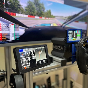 May include: A black racing simulator setup with a large screen displaying a racing game, a steering wheel, and a dashboard with a digital display showing the lap time and speed. The dashboard also shows the gap between the driver and the car in front.
