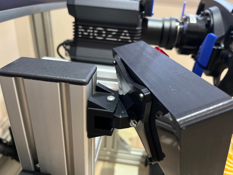 May include: Close-up of a black metal bracket attached to a silver aluminum rail. The bracket has a screw and a small black plastic piece. The bracket is attached to a black metal frame.
