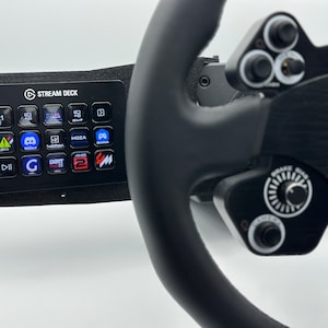 May include: A black racing wheel with a black Stream Deck mounted to the left side. The Stream Deck has a variety of buttons with different logos and icons. The racing wheel has a black leather grip and a silver knob on the right side. The text "MOZA" is visible on the wheel.