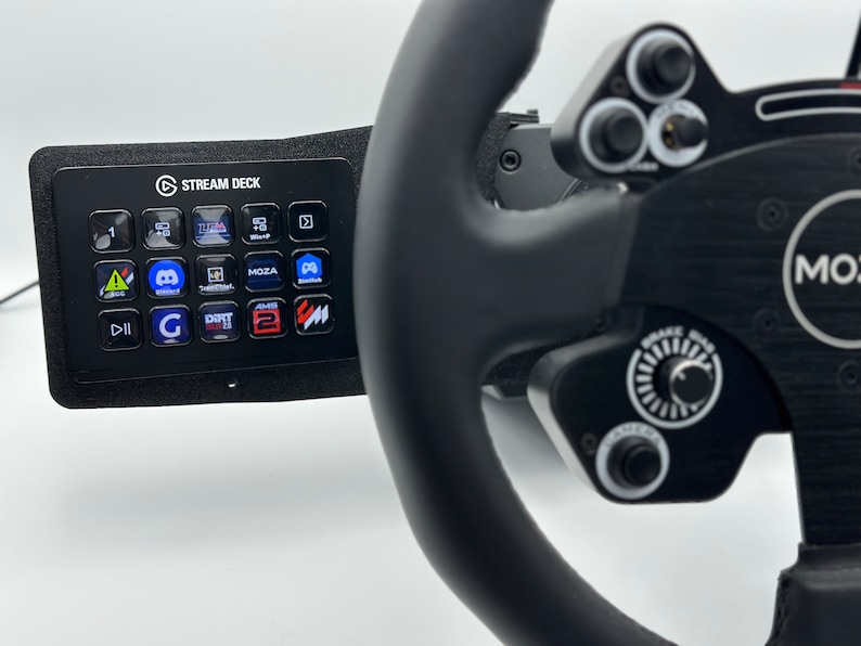 May include: A black racing wheel with a black Stream Deck mounted to the left side. The Stream Deck has a variety of buttons with different logos and icons. The racing wheel has a black leather grip and a silver knob on the right side. The text "MOZA" is visible on the wheel.