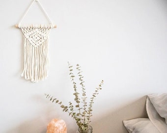 May include: A white macrame wall hanging with a wooden dowel, a salt lamp, a glass jar with dried eucalyptus branches, and a white candle on a wooden table.