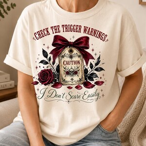 Può includere: Una t-shirt color crema con una stampa grafica di un fiocco rosso scuro legato a un'etichetta in stile vintage con la scritta CAUTION e l'illustrazione di un teschio. Il design include rose rosso scuro, foglie nere e il testo CHECK THE TRIGGER WARNINGS e I Don't Scare Easily.