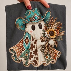 Op de afbeelding: Een donkergrijs t-shirt met een afbeelding van een geest die een turquoise cowboyhoed met een zilveren concho draagt. De geest houdt een boeket zonnebloemen en tarwe vast, gestrikt met een luipaardprint, op een koeienhuid patchwork patroon.