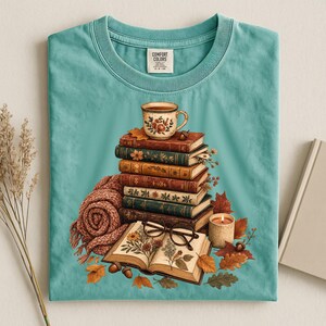 Op de afbeelding: Een blauwgroen t-shirt met ronde hals en een afbeelding van een stapel van vijf oude boeken met daarop een bloemenmok. Een open boek met botanische illustraties, een leesbril, een bruine gebreide sjaal, een kaars en eikels liggen eromheen.