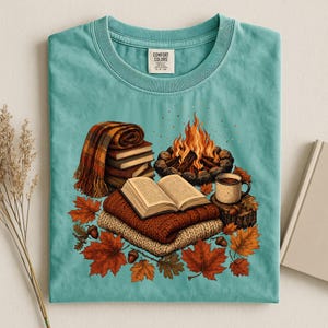 May include: A teal t-shirt with a graphic print featuring a campfire, a stack of books, an open book resting on folded sweaters, a mug, and scattered autumn leaves. The inner neck label displays the text Comfort Colors in black lettering.