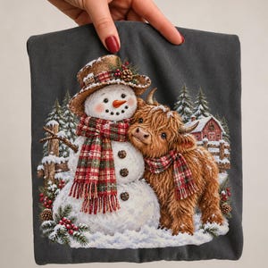 May include: A dark grey t-shirt featuring a graphic print of a snowman wearing a brown hat and a red and green plaid scarf, standing next to a fluffy brown Highland cow wearing a matching plaid scarf, with a snowy barn and pine trees.
