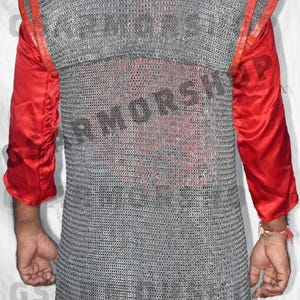 May include: A silver chainmail shirt with red leather trim. The shirt is being worn by a person, but their face is not visible. The shirt is made of interlocking metal rings.