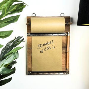 May include: A wooden wall-mounted notepad with a roll of paper at the top and a sheet below. The sheet reads "50 metres of lists" in black ink. A pencil is attached to the side.