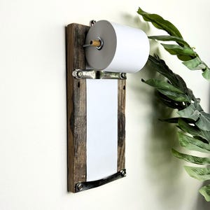 May include: A rustic wooden toilet roll holder with an integrated notepad. It features a roll of white toilet paper and a white notepad, secured with metal hardware. The wood has a weathered appearance, enhancing its rustic charm.
