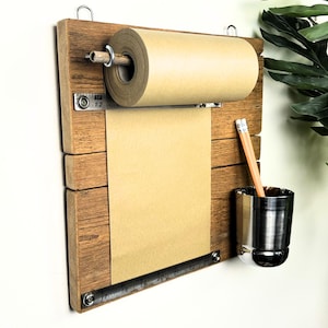 May include: A wooden wall-mounted paper roll holder with a roll of brown paper. It includes a metal pencil cup and a pencil. The holder is made from reclaimed wood, offering a rustic aesthetic. Ideal for notes and lists, it adds a vintage touch.