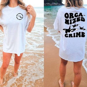 May include: White t-shirt with a black graphic on the front that says "Sea-Sized Crime" and a black graphic on the back that says "ORCA-NIZED CRIME" with a group of orcas swimming.