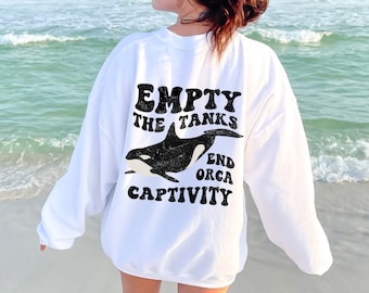 Może przedstawiać: Biała bluza z czarnym nadrukiem graficznym przedstawiającym orkę i tekstem "Empty the Tanks End Orca Captivity".