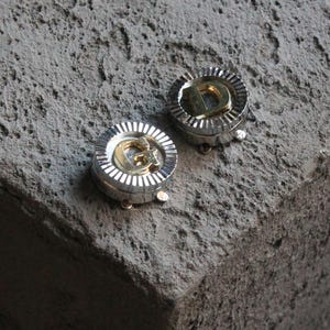 Pode incluir: Dois botões de punho de metal em tom prateado e dourado com uma letra 'D' em cada um. Os botões de punho estão sobre uma superfície cinzenta texturizada.