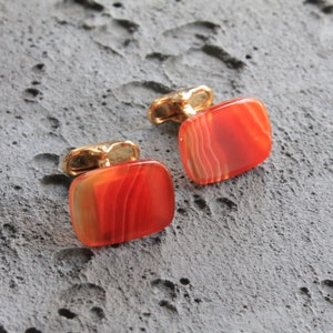 Puede incluir: Un par de gemelos con inserciones de piedra de ágata naranja. Los gemelos tienen un acabado dorado.