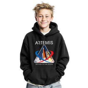 Puede incluir: Sudadera con capucha negra con la palabra "ARTEMIS" en blanco sobre un gráfico triangular con un cohete y el texto "RETURN TO THE MOON" en blanco. El gráfico tiene detalles en rojo y azul.
