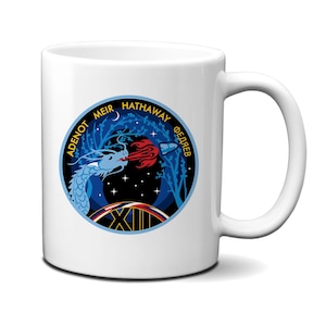 Puede incluir: Taza de cerámica blanca con un diseño circular que representa un dragón azul que lanza fuego a un cohete. El diseño incluye texto en inglés y ruso, con un fondo de cielo estrellado. La taza tiene un asa curvada.