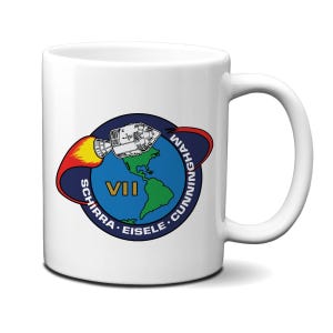 May include: White ceramic mug with a blue, red, yellow, and green graphic of a space capsule orbiting Earth. The text "VII SCHIRRA. EISELE. CUNNINGHAM" is printed around the graphic.