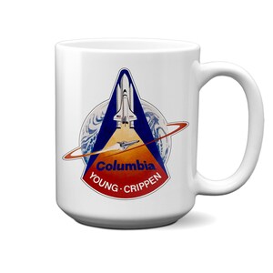 Könnte beinhalten: Ein weißer Keramikbecher mit einer Grafik des Missionsabzeichens der Space Shuttle Columbia. Das Design zeigt ein blau-oranges dreieckiges Emblem mit einem Shuttle, dem Text Columbia sowie den Namen Young und Crippen vor einem Globus.
