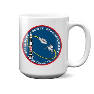 May include: White ceramic mug with a blue and yellow Apollo IX mission patch featuring a rocket, lunar module, and the names of the astronauts: McDivitt, Scott, and Schweickart.