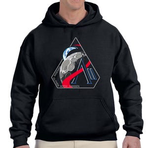 Könnte beinhalten: Schwarzer Kapuzenpullover mit einer weißen, roten und blauen Grafik des Mondes, der Erde und dem Buchstaben "A" in einem Dreieck. Die Namen "WISEMAN", "GLOVER", "KOCH" und "HANSEN" sind aufgedruckt.