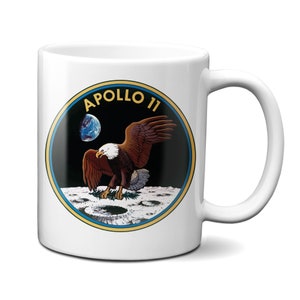 Pode incluir: Caneca de café de cerâmica branca com uma imagem em preto e branco do patch da missão Apollo 11. O patch mostra uma águia pousando na lua com a Terra ao fundo. O texto "APOLLO 11" é impresso em amarelo acima da águia.
