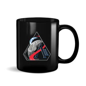 May include: Black ceramic mug featuring a triangular design with a stylized moon, Earth, and the letter "A". The design includes red and blue accents, with the names "WISEMAN", "GLOVER", "KOCH", and "HANSEN" printed on the mug.