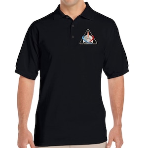 Könnte beinhalten: Ein schwarzes Poloshirt mit einem kleinen gestickten Aufnäher auf der linken Brust. Der Aufnäher zeigt eine Rakete in einem Dreieck mit einem blauen und roten Kreis darum.