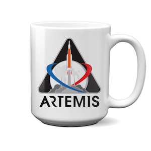 Könnte beinhalten: Weißer Keramikbecher mit dem Logo des Artemis-Programms. Das Logo zeigt eine Rakete, die vor einem Vollmond startet, mit blauen und roten Schwüngen. Das Wort "ARTEMIS" ist darunter in Schwarz gedruckt.