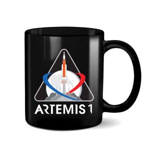 Peut inclure: Mug en céramique noire avec un graphique blanc d'une fusée lancée vers la lune avec une orbite rouge et bleue autour d'elle. Le graphique est à l'intérieur d'un triangle blanc avec le texte "ARTEMIS 1" en dessous.