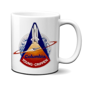 Könnte beinhalten: Ein weißer Keramikbecher mit dem STS-1 Missionsabzeichen. Das Design zeigt ein Space Shuttle, eine stilisierte Erde sowie den Text Columbia, Young und Crippen. Die Grafik ist mittig auf der Vorderseite des Bechers platziert, mit einem Henkel rechts.