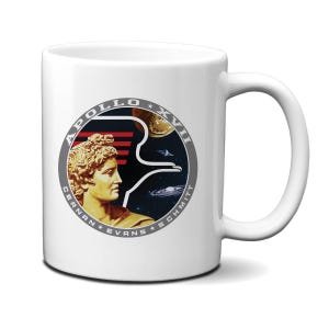 Könnte beinhalten: Weiße Keramiktasse mit einem Schwarzweißbild des Apollo-17-Missionsabzeichens. Das Abzeichen zeigt ein stilisiertes Bild des griechischen Gottes Apollon, einen zunehmenden Mond und eine Galaxie. Der Text "Apollo XVII" ist in einem Kreis um das Bild gedruckt. Die Namen der Astronauten, "Cernan, Evans, Schmitt", sind in einem Kreis unter dem Bild gedruckt.