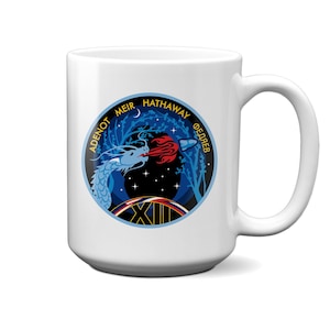 Puede incluir: Taza de cerámica blanca con un diseño circular. El diseño presenta un dragón azul, una llama roja y el texto "ADENOT MEIR HATHAWAY ФЕДЯЕВ". El diseño incluye un cielo nocturno y el número romano XII. La taza tiene un asa grande.