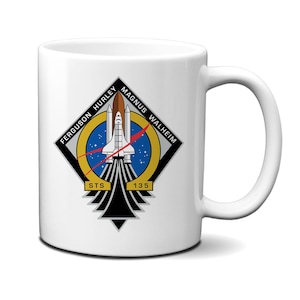 May include: A white ceramic mug featuring the NASA STS-135 mission patch. The diamond-shaped emblem displays a space shuttle launching into a blue starry sky, with the text FERGUSON HURLEY MAGNUS WALHEIM and STS 135 printed in black and gold.