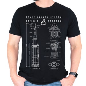Może przedstawiać: Czarny T-shirt z białym nadrukiem graficznym przedstawiającym rakietę Space Launch System (SLS) i statek kosmiczny Orion. Grafika zawiera tekst "Space Launch System Artemis Program".