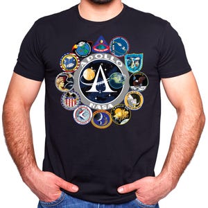 May include: Navy blue t-shirt with a circular design featuring the words "APOLLO" and "NASA". The design includes mission patches and a central "A" symbol. The shirt has a crew neck.
