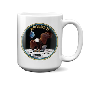 May include: White ceramic mug with a black and white image of an eagle landing on the moon. The image is surrounded by a yellow circle with the text "APOLLO 11" in yellow.