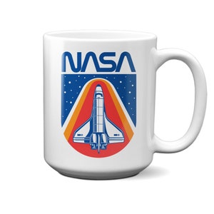 May include: White ceramic mug with a blue and red NASA logo featuring a space shuttle against a starry sky background.