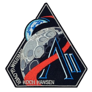 May include: A triangular patch with a design featuring a grey moon, a blue and white planet, and a large letter "A". The patch includes the names GLOVER, WISEMAN, KOCH, and HANSEN. Red and blue accents are also present.
