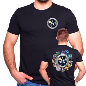 May include: Two black t-shirts with NASA Apollo mission patches. The shirt on the left has a single patch with the Apollo logo and NASA text. The shirt on the right has a larger patch with the Apollo logo, NASA text, and a circle of smaller patches around it.