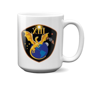 Puede incluir: Una taza de cerámica blanca con un gráfico en forma de escudo. El diseño muestra un dragón amarillo sobre una Tierra azul, con el número romano XIII, un sol, estrellas, una luna y una estación espacial sobre un fondo negro.