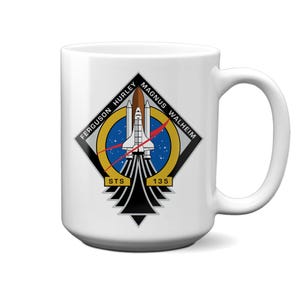 May include: A white ceramic mug featuring the STS-135 mission patch. The design includes a space shuttle launching into a blue circular field with stars, surrounded by a yellow border. Text reads FERGUSON HURLEY MAGNUS WALHEIM and STS 135.
