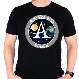 Könnte beinhalten: Schwarzes T-Shirt mit einem weißen NASA Apollo-Missions-Patch, der ein stilisiertes 'A' mit Mond und Erde im Hintergrund zeigt.